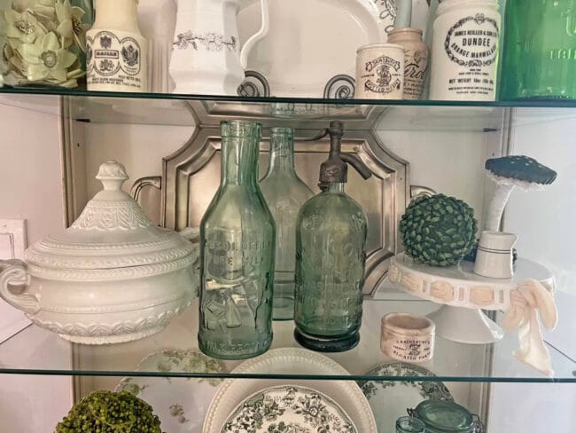 glass shelves in hutch with green bottles, white tureen, silver tray and cake plate with green ball and mushroom on top