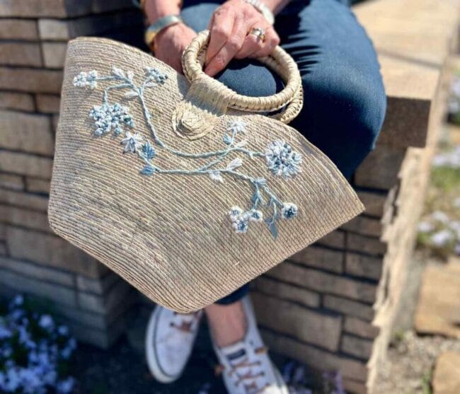 A lady in jeans holding a straw purse with blue embroidery