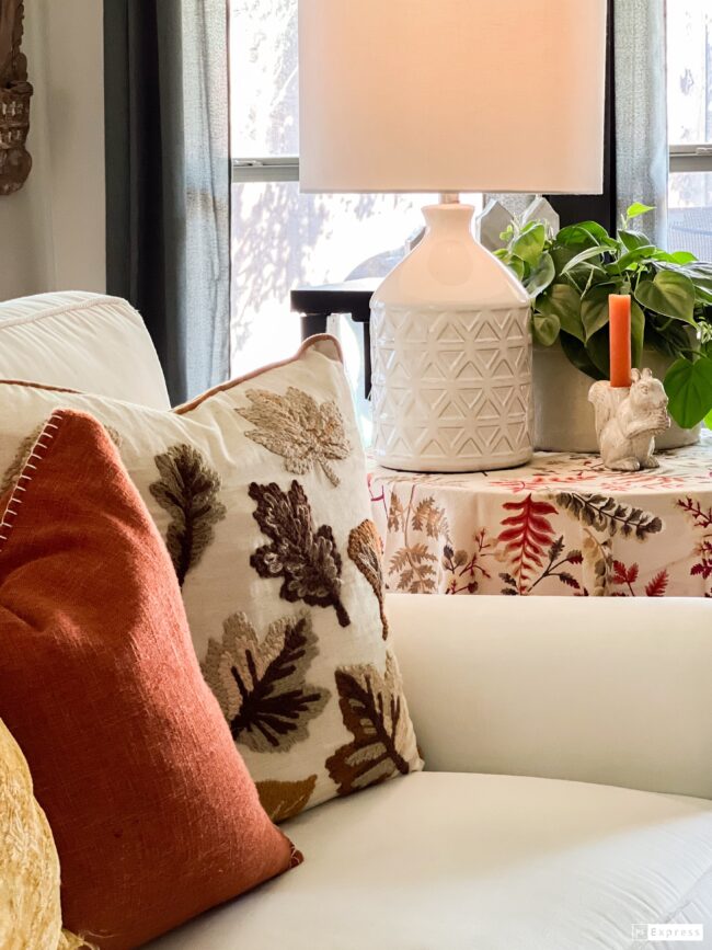 white sofa with fall leaf and orange pillows with a side table with a white lamp and squirrel candle next to a plant