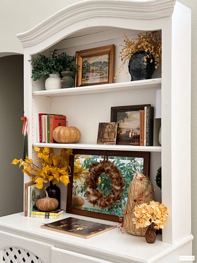 close up of hutch shelves decorated with books, a mirror with a feather wreath and faux stems