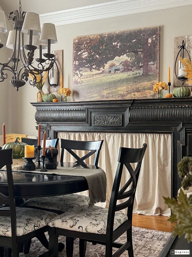 black dining table with black mantel on wall with large farm print on wall and golden candles and flowers.