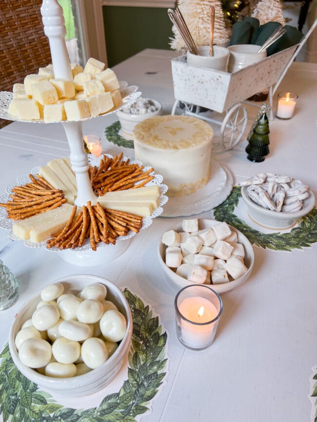 dessert table with white candies in white bowls, a white cake and a 2-tiered plate rack with pretzels and cubed cheese