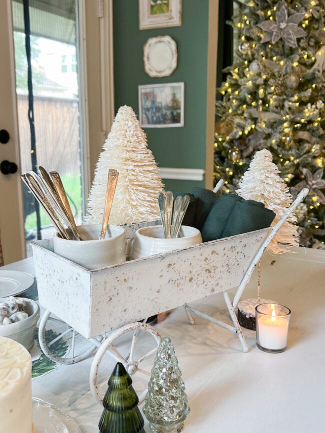 Small white wheelbarrow sitting on table with silverware, green napkins and a candle in corner