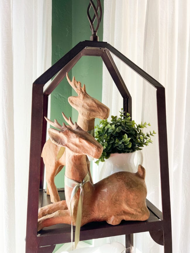 2 paper mache deer with green ribbon and a white pot of greenery on the top rack of the plate stand