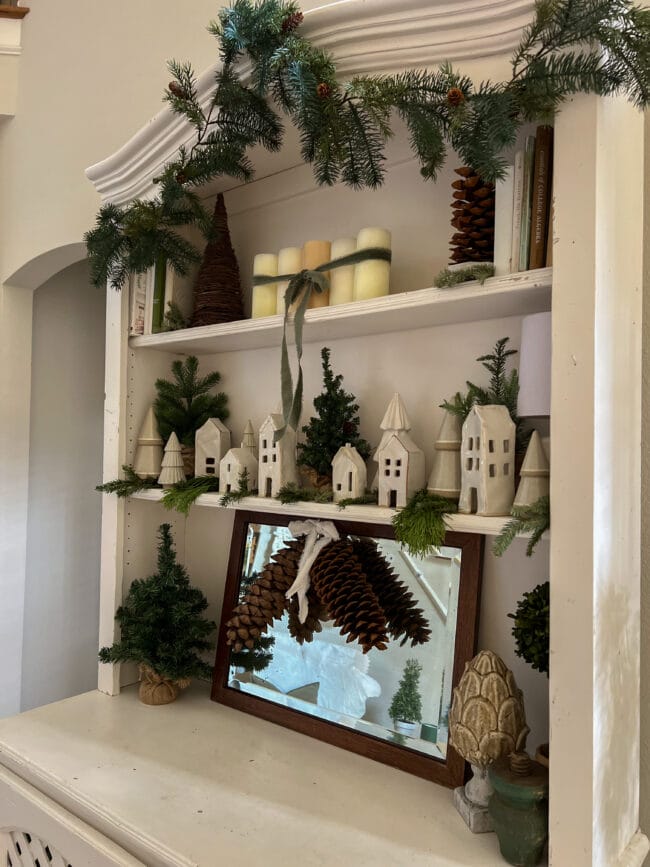 white hutch shelves with white houses, live greenery stems and books and a large mirror on the bottom with pinecones.