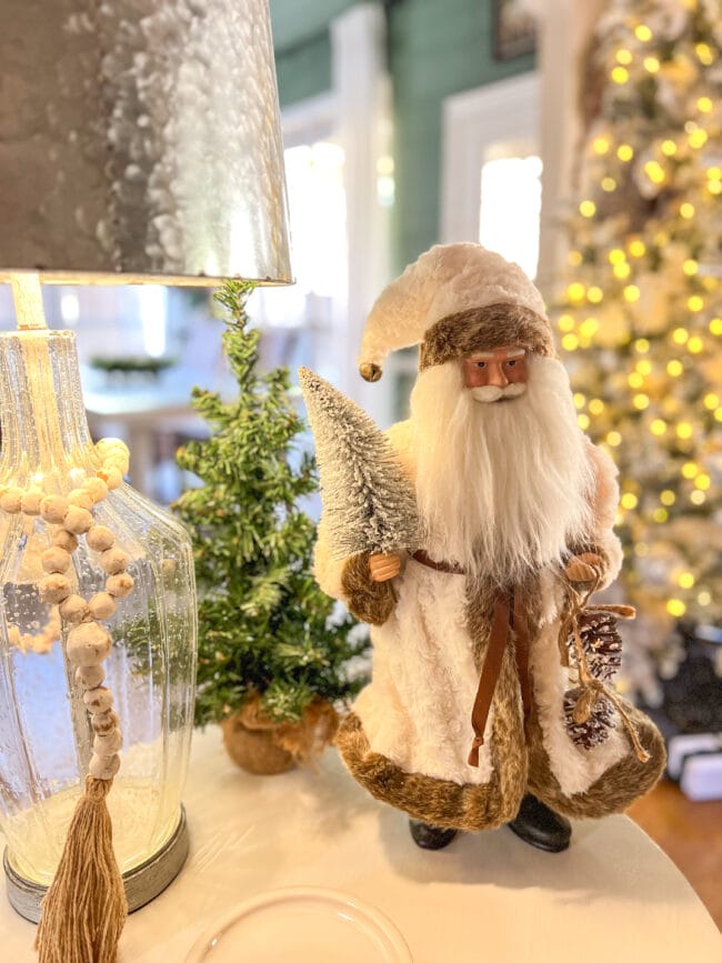 White fuzzy coat on Santa, small tree and clear glass lamp on white table