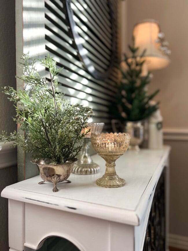 white table with antique mercury glass candy dishes with candles inside and faux greenery stem in a vintage silver creamer