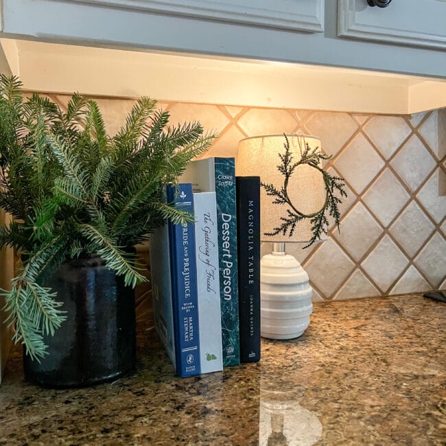 black crock with live greenery, cookbooks and lamp with tiny wreath hanging on shade.