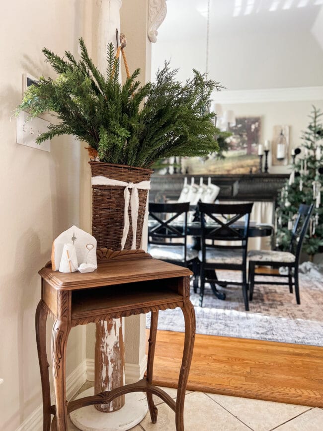 Small wooden entry table with a manger scene and a hanging basket of green stems with the dining room in the background.