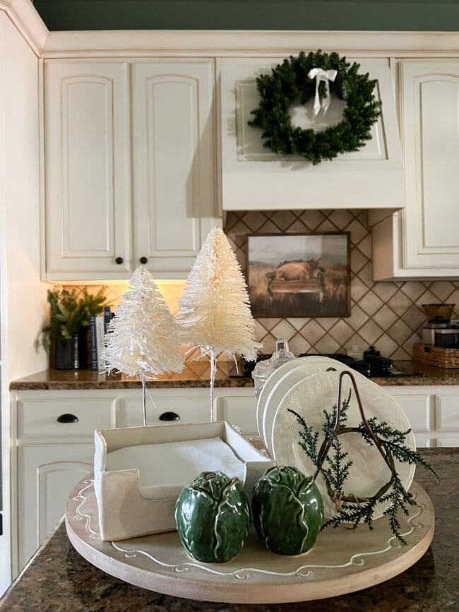 kitchen island with white bottle brush trees, stacked plates, and black crock with live greenery in the back ground.