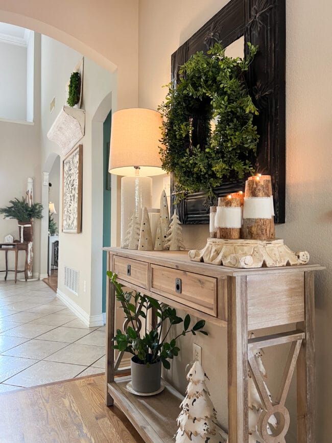 light wood console table with white ceramic and metal trees, a wreath on the black mirror and several handmade limb candles