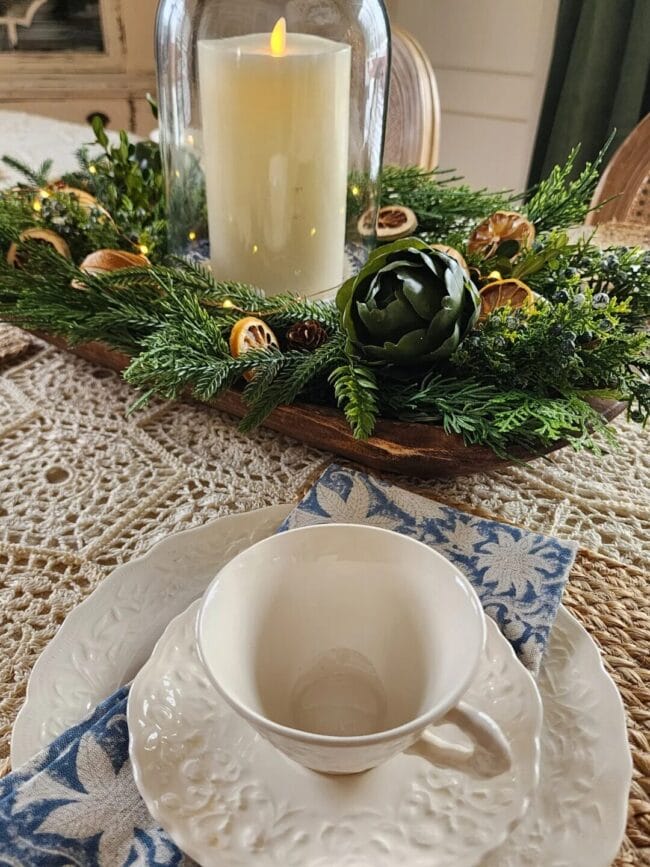 dining table with greens, oranges and a hurricane over a candle in a dough bowl with a place setting of antique plates and a blue napkin