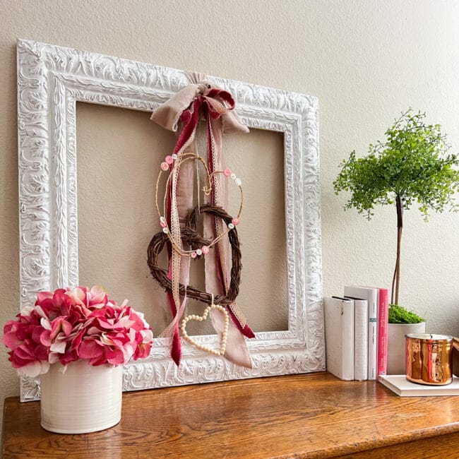 white frame with Valentine's day wreaths and ribbons hanging inside and sitting on a wooden chest with pink flowers, books and a topiary.