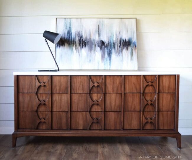 dark stained dresser with black lamp sitting on top and an abstract painting on the wall