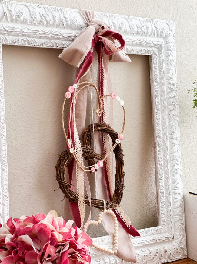 white frame leaning against the wall with 3 heart wreaths hanging inside. Has pink ribbons too.