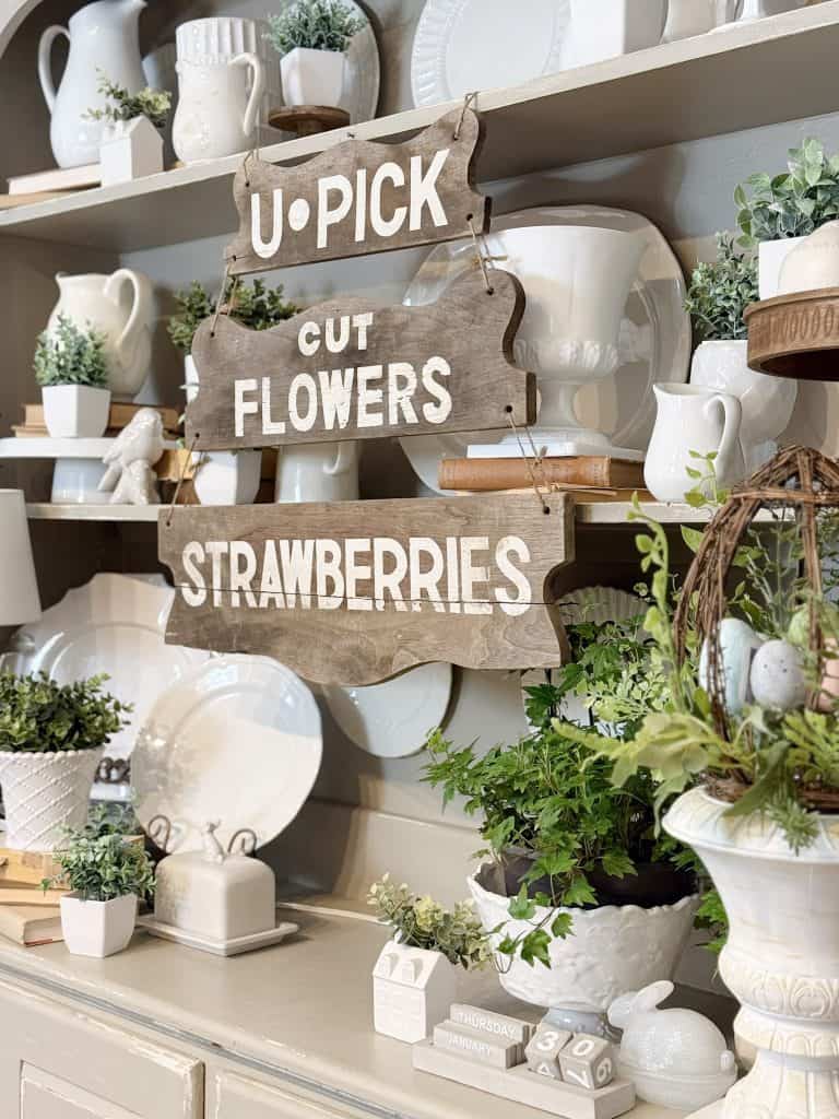 hutch shelves filled with greenery, white plates and a wooden spring sign hanging on the front