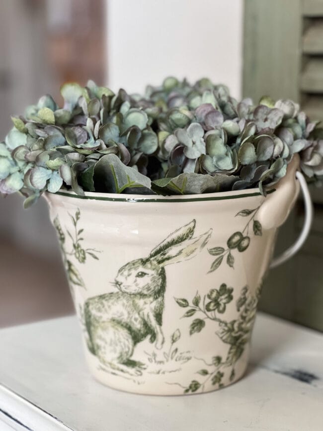 green and white ceramic bucket with a rabbit on the side and filled with faux hydrangeas.