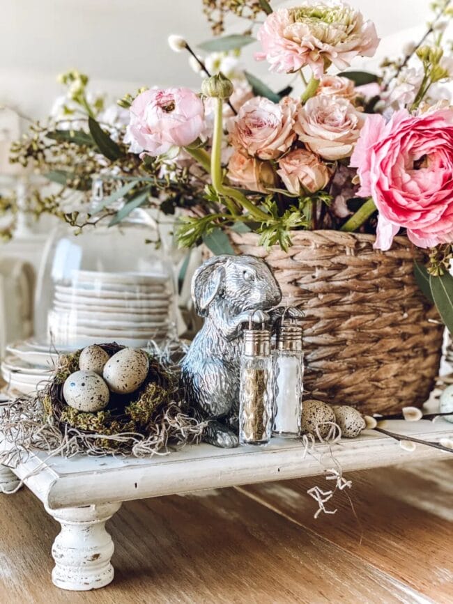 white stand with bunny, nest with eggs, a stack of white dishes under a cloche with a basket of flowers