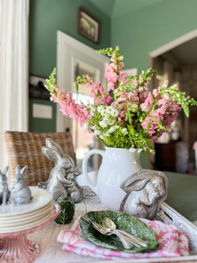 table centerpiece with pink and white flowers in a white pitcher, silver bunnies and a stack of plates with tiny bunnies on top