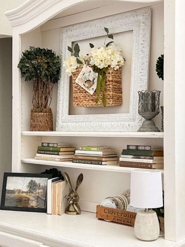 Close up of hutch with topiaries, basket of flowers, books, plates and brass bunny head next to a pastoral photo