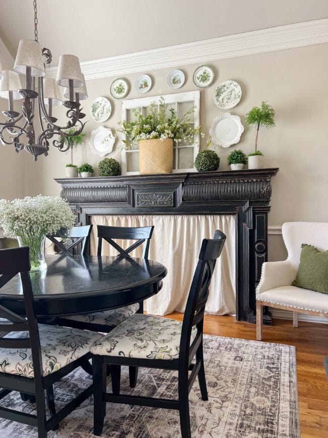 dining room with black table and mantel. Mantel is decorated with green and white plates and lots of greenery balls and topiaries and spring flowers.