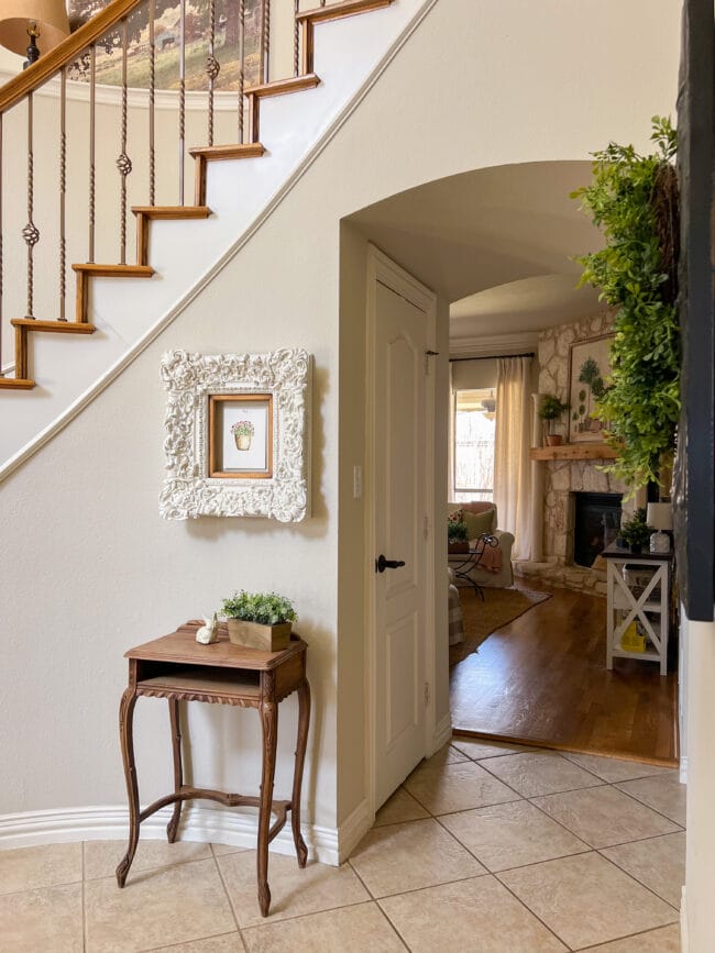 small vintage wooden table with white ornate frame on wall above it and view of living room and stone fireplace