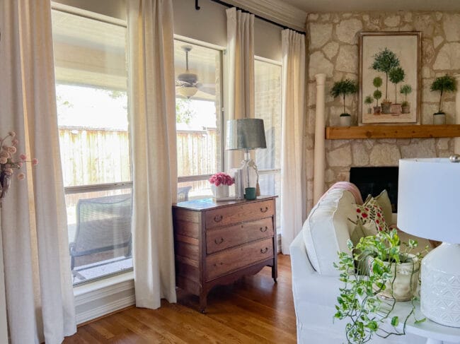 living room windows with cream curtains, an antique 3 drawer chest and accessories