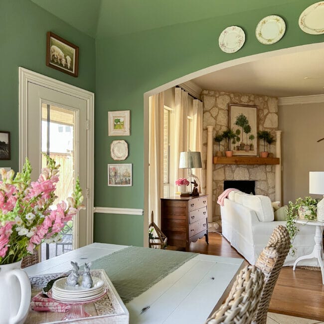 green walls in the breakfast area with plates and prints on the walls, pink flowers and the family room in the background 